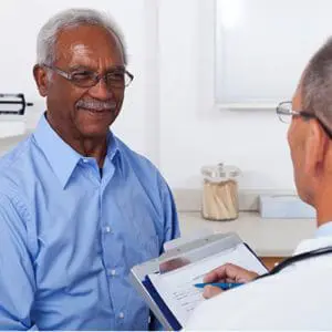 A mature male doctor writing on a clipboard while talking to a smiling male senior patient in the doctor's office.