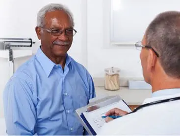 A mature male doctor writing on a clipboard while talking to a smiling male senior patient in the doctor's office.