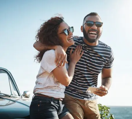 A young couple both wearing sunglasses outside while sitting on the hood of their car smiling.