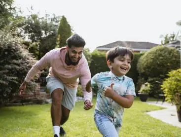 A young man playing chase with his son in the garden outside both smiling.