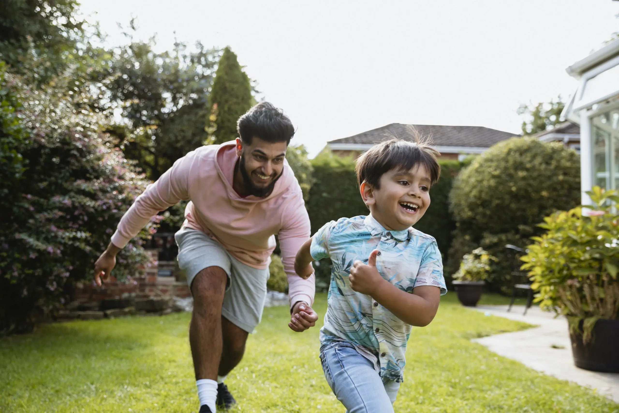 A young man playing chase with his son in the garden outside both smiling.