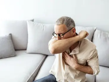 A mature male coughing into his elbow while sitting on a couch.