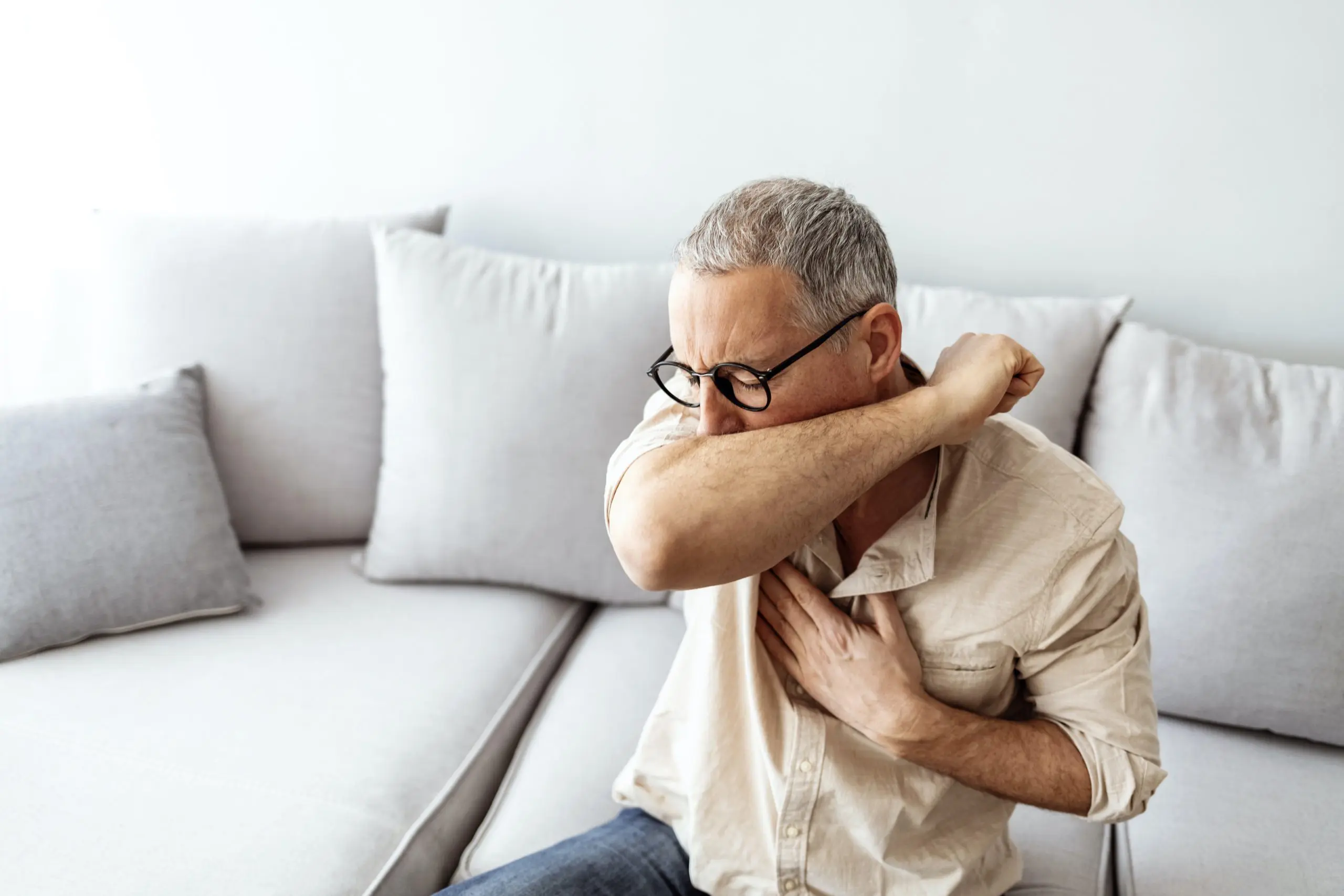 A mature male coughing into his elbow while sitting on a couch.