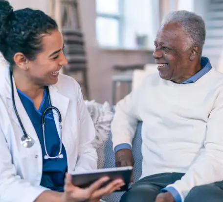 A female doctor holding a digital tablet looking at a senior male patient both sitting on a couch at the patient's home smiling.