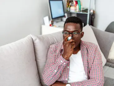A young male using an inhaler while sitting on his couch at home.
