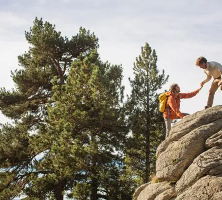 A male helping another male wearing a backpack and orange jacket climb up a rock with pine trees in the background.