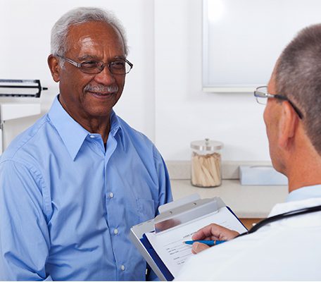A mature male doctor writing on a clipboard while talking to a smiling male senior patient in the doctor's office.