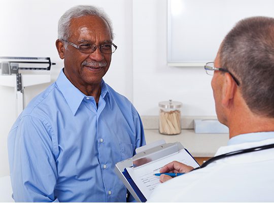 A mature male doctor writing on a clipboard while talking to a smiling male senior patient in the doctor's office.