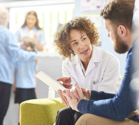 A female pharmacist sitting with a male patient in a pharmacy discussing his prescription. In the background a senior woman and granddaughter stand at the dispensing counter and are served by a different female pharmacist..