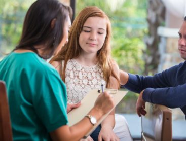 A female nurse writing on a clipboard helping treat a young female teen with her allergies.