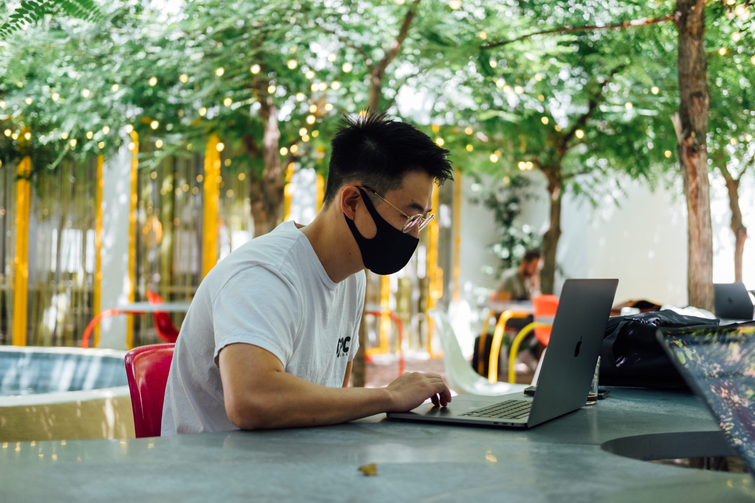 A young male sitting outside at a table wearing a black face mask working on his computer, surrounded by a few trees with lights on them.