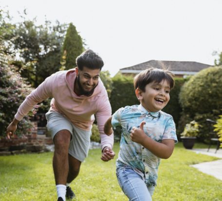 A young man playing chase with his son in the garden outside both smiling.