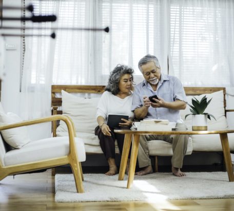 A senior male looking down at his phone while showing his senior wife both sitting on the couch in their living room.