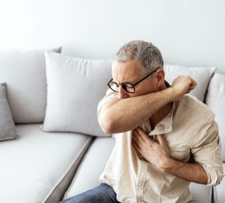 A mature male coughing into his elbow while sitting on a couch.