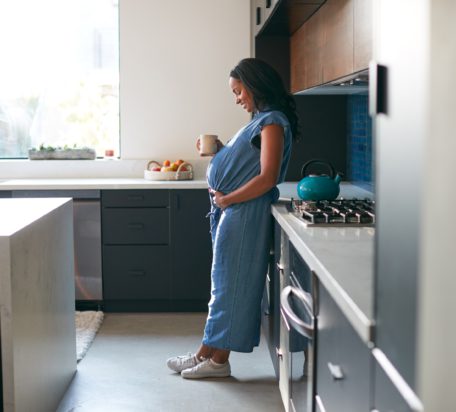 A pregnant woman standing in her kitchen at home smiling while looking down at her stomach with a hot drink in her hand.