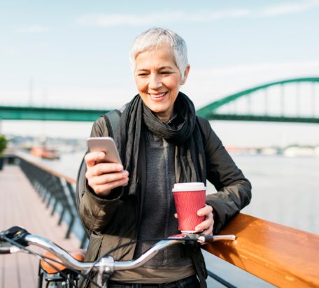 A senior woman standing next to her bike by a fence, smiling while typing a message on the phone and drinking coffee outside by a river.