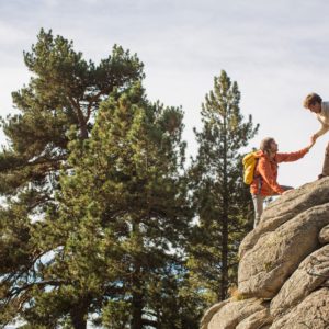 A male helping another male wearing a backpack and orange jacket climb up a rock with pine trees in the background.